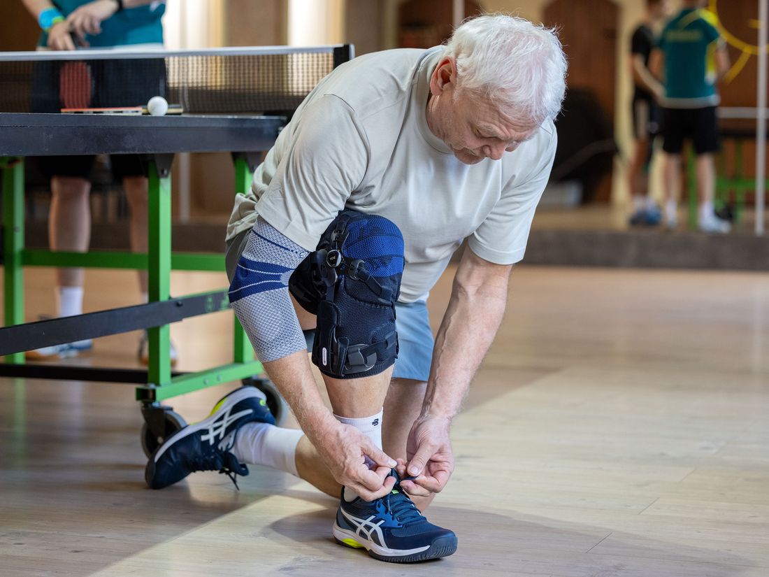 Älterer Sportler mit Knieorthese und Kompressionsbandage bindet vor dem Tischtennisspiel seinen Schuh